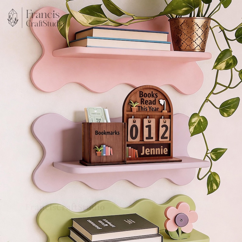 May include: Three-tiered, wavy-edged shelves in pink and green hold books, a potted plant, and decorative wooden accessories. A wooden calendar displays "Books Read This Year" and the number "012" with the name "Jennie". A wooden box reads "Bookmarks".