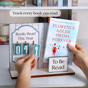 May include: A wooden book tracker with the text "Books Read This Year" and the numbers "041". A book titled "Florence Adler Swims Forever" is displayed next to a "To Be Read" sign. The text "Track every book you read" is also visible.