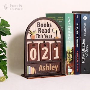 Op de afbeelding: Een houten boekenplankdecoratiestuk met de tekst "Books Read This Year" en de cijfers "021". De naam "Ashley" wordt onder de cijfers weergegeven. De decoratie bevat kleine illustraties van een plant en boeken.