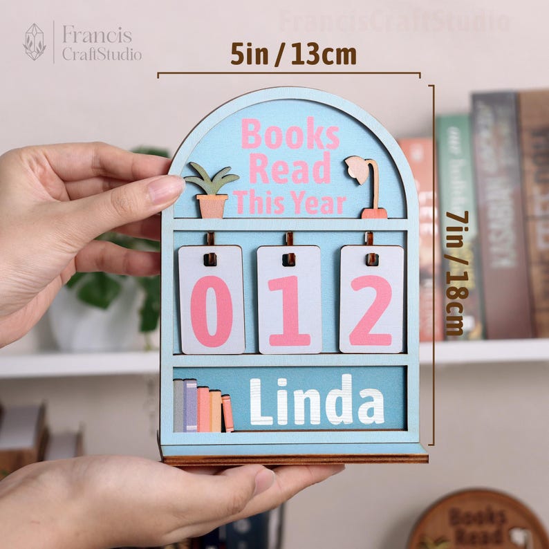 Puede incluir: Un rastreador de libros de madera azul claro con el texto "Books Read This Year" y el nombre "Linda". El rastreador mide 13 cm de ancho y 18 cm de alto. Tiene tres ranuras num&eacute;ricas que muestran "012".