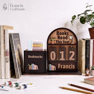 May include: A wooden desk organizer with a bookmark holder and a counter that reads "Books Read This Year: 012". The organizer has a dark brown finish with the name "Francis" at the bottom. A small plant and a lamp are also featured.