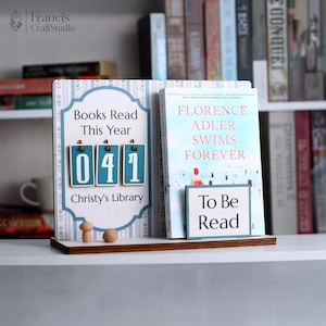May include: A wooden book stand with a blue and white design, displaying a book and a counter that reads "041 Books Read This Year". The book is titled "Florence Adler Swims Forever". A small sign reads "To Be Read".