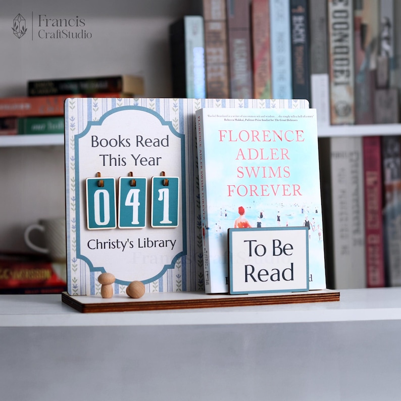 May include: A wooden book stand with a blue and white design, displaying a book and a counter that reads "041 Books Read This Year". The book is titled "Florence Adler Swims Forever". A small sign reads "To Be Read".