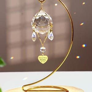 May include: A gold metal stand with a crystal ball ornament hanging from it. The ornament has a heart-shaped charm with the inscription "In Loving Memory Grandma 1937-2023".