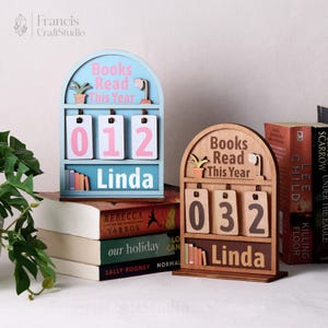 Puede incluir: Dos contadores de libros de madera. Uno es azul claro con n&uacute;meros rosas y el texto "Books Read This Year" y "Linda". El otro es de color madera con n&uacute;meros marrones y el mismo texto. Ambos tienen una estanter&iacute;a con libros en miniatura.