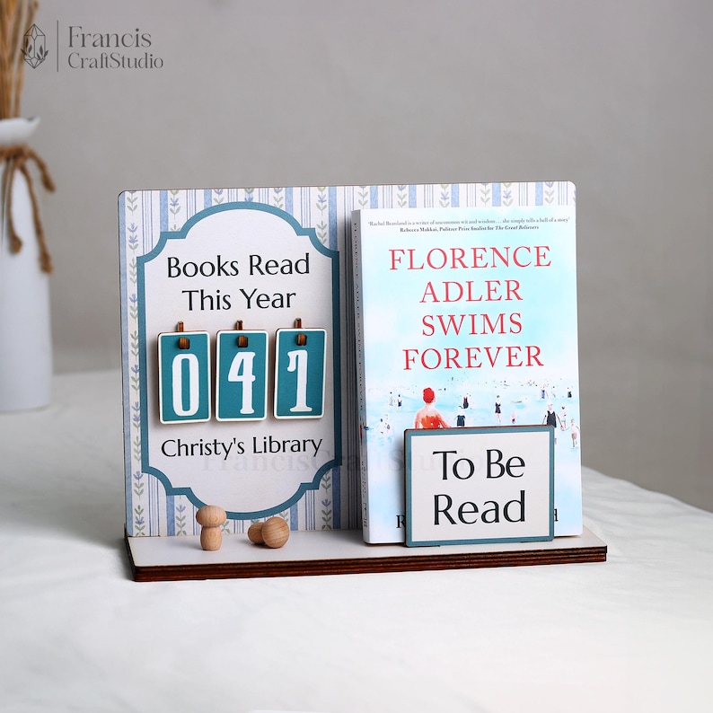 May include: A wooden book display with the text "Books Read This Year" and the number "041". It also features a book titled "Florence Adler Swims Forever" and a "To Be Read" sign. The display is on a white surface.