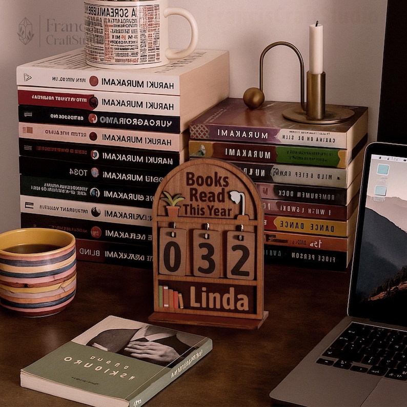 Puede incluir: Un escritorio con libros, una taza y un candelabro. Un calendario de madera muestra "Books Read This Year", "032" y "Linda". Una computadora port&aacute;til est&aacute; abierta. La escena sugiere un ambiente de lectura y trabajo.