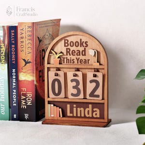 May include: Wooden book counter with the text "Books Read This Year" and the number "032". The counter has a small shelf with miniature books and the name "Linda". Several books are visible in the background.