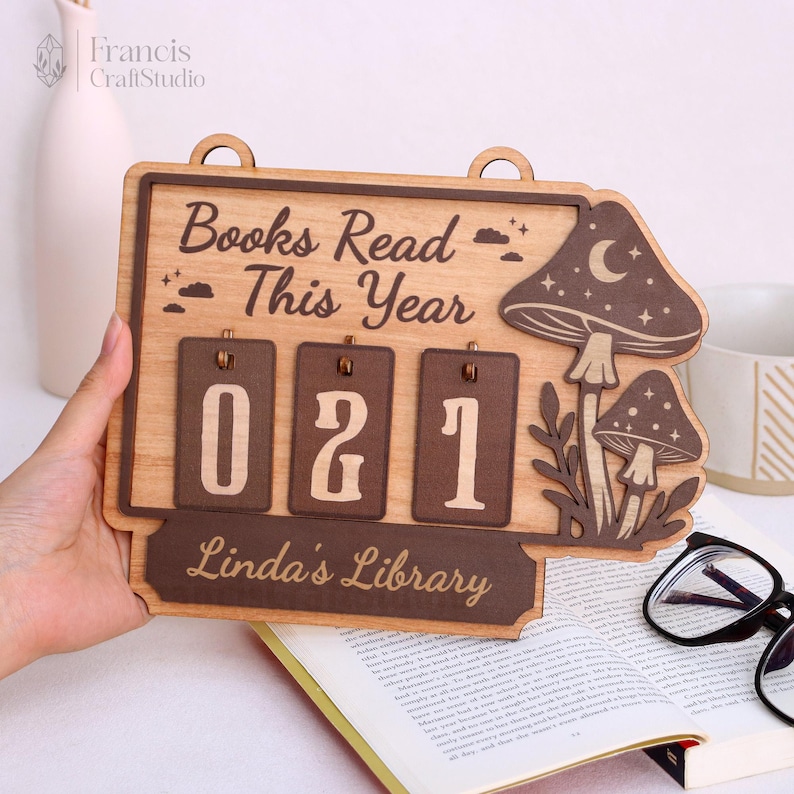 以下が含まれることがあります： 「Books Read This Year」の文字と数字「021」が入った木製のブックカウンター。カウンターにはキノコの模様と「Linda's Library」の文字が入っています。