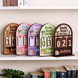May include: Five wooden book trackers in various colors, each with a different number displayed. The trackers have the text "Books Read This Year" and a space to write a name. The numbers are displayed on a flip-style calendar.