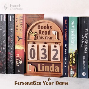 Puede incluir: Un elemento decorativo de estanter&iacute;a de madera con el texto "Books Read This Year" y el n&uacute;mero "032". El nombre "Linda" se muestra debajo de los n&uacute;meros. El texto "Personaliza tu nombre" est&aacute; en la parte inferior.