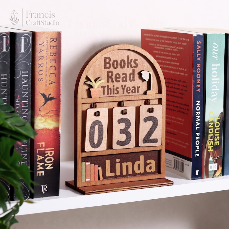 Puede incluir: Un rastreador de libros de madera con el texto "Books Read This Year" y el n&uacute;mero "032". El rastreador tiene el nombre "Linda" y peque&ntilde;as ilustraciones de una planta y libros. Estanter&iacute;as en el fondo.