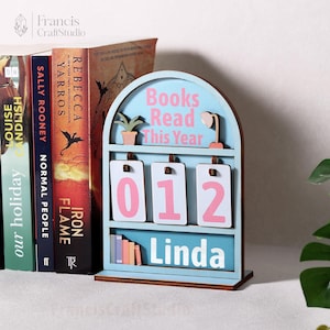 Puede incluir: Un mostrador de libros de madera azul claro con el texto "Books Read This Year" y los n&uacute;meros "012" en rosa. El nombre "Linda" se muestra en la parte inferior. Hay una peque&ntilde;a planta y una l&aacute;mpara en la parte superior.