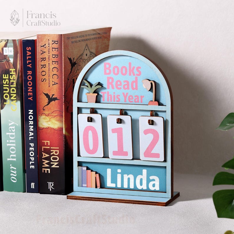 Puede incluir: Un mostrador de libros de madera azul claro con el texto "Books Read This Year" y los n&uacute;meros "012" en rosa. El nombre "Linda" se muestra en la parte inferior. Hay una peque&ntilde;a planta y una l&aacute;mpara en la parte superior.