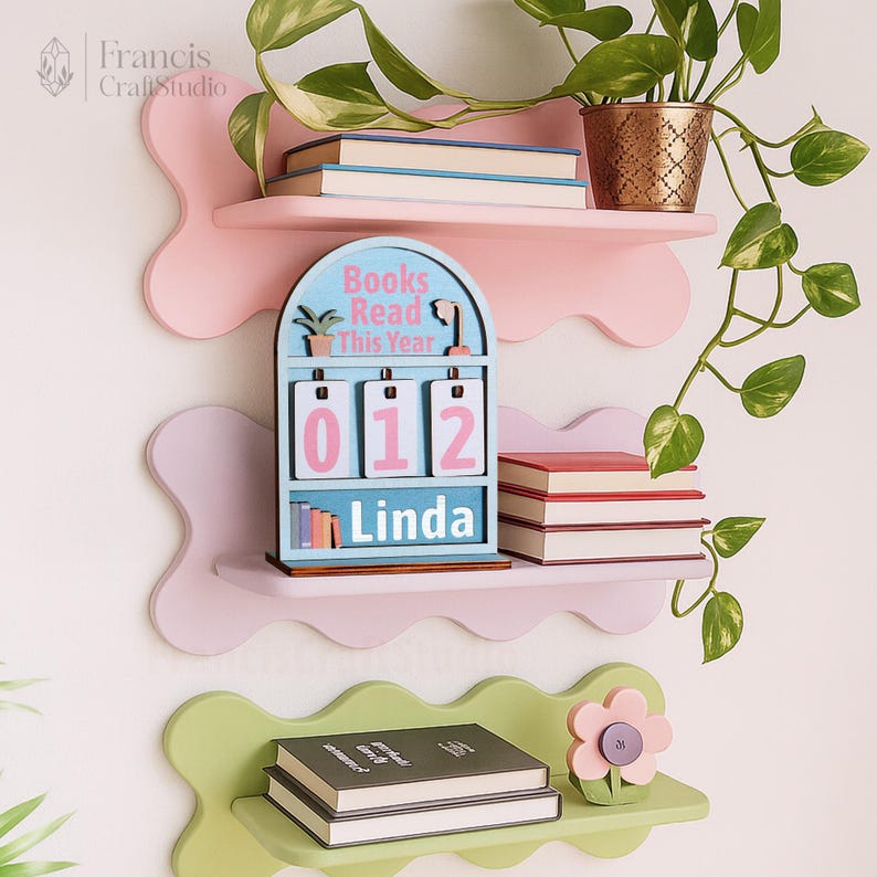 Puede incluir: Un conjunto de estanter&iacute;as de pared en tonos pastel rosa y verde con tres niveles. Contiene libros, una decoraci&oacute;n floral y un calendario de madera que dice "Books Read This Year" con el nombre "Linda" debajo de los n&uacute;meros.