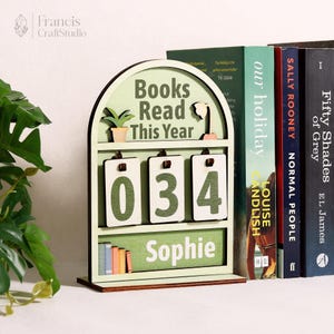 May include: A wooden book tracker with the text "Books Read This Year" and the number "034". The tracker has a light green arched frame with a shelf and a personalized nameplate that says "Sophie". Several books are visible in the background.