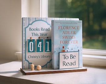 May include: A wooden bookshelf with two books. One book has the text "Books Read This Year" and displays the number "041". The other book is titled "Florence Adler Swims Forever" with a "To Be Read" sign. Two small wooden figures are on the shelf.