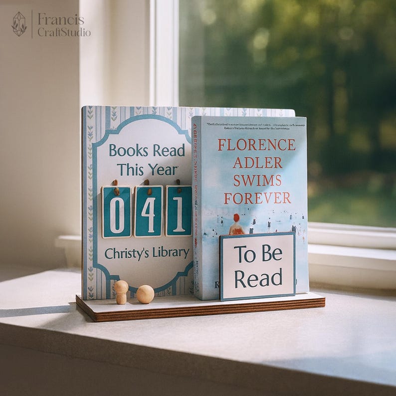 May include: A wooden bookshelf with two books. One book has the text "Books Read This Year" and the number "041". The other book has the text "FLORENCE ADLER SWIMS FOREVER" and "To Be Read". The bookshelf is on a windowsill.