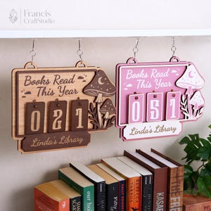 以下が含まれることがあります： 「Books Read This Year」と「Linda's Library」の文字が入った2つの木製ブックトラッカー。1つは薄茶色で、濃い茶色の数字とキノコのアクセントがあり、もう1つはピンク色で、白い数字とキノコのアクセントがあります。どちらも吊り下げられています。