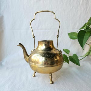May include: A vintage brass teapot with a curved spout, a decorative handle, and three small legs. The teapot has etched floral patterns and a polished finish. It is a decorative item, likely for display.