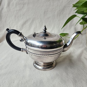 Peut inclure: Une théière en argent avec une poignée noire et un couvercle décoratif. La théière a un corps rond avec un motif en forme de corde autour du milieu et repose sur une base ronde. Le bec verseur est courbé.