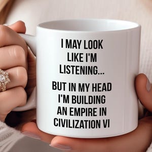 Op de afbeelding: Een witte keramische koffiemok met zwarte tekst die luidt: "Ik mag eruit zien alsof ik luister... maar in mijn hoofd bouw ik een imperium in Civilization VI."