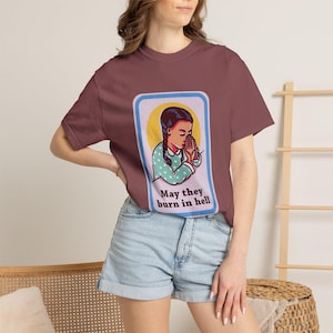 Può includere: Una t-shirt a maniche corte color marrone con una grafica rettangolare di una persona con capelli intrecciati in una camicia a pois color verde acqua, con le mani giunte in preghiera su un cerchio giallo. Il testo sotto recita, May they burn in hell.