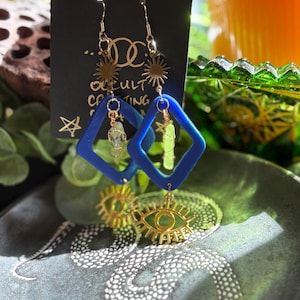 May include: A pair of dangle earrings with gold-colored hooks and accents. Each earring features a blue diamond-shaped frame, a gold sunburst, and a gold eye charm. The earrings hang from a black card with the text "occult creating".