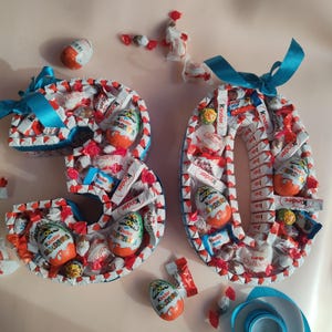 Könnte beinhalten: Zwei Pappzahlen, 3 und 0, verziert mit verschiedenen Schokoladenbonbons, darunter Kinder Überraschungseier, Kinder Schokoladentafeln und Raffaello Pralinen, die mit einem blauen Band zusammengebunden sind.