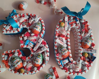 May include: A blue ribbon tied around two cardboard numbers, 3 and 0, decorated with various chocolate candies, including Kinder Surprise eggs, Kinder chocolate bars, and Raffaello chocolates.