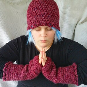 May include: A burgundy crochet hat and matching fingerless gloves. The hat has a scalloped brim and the gloves have a ruffled edge. The items are made of a textured yarn and appear handmade. The person in the image is wearing a black top.