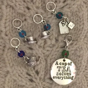 May include: A selection of silver knitting stitch markers with tea-themed charms. Charms include a teapot, teacups, and a tea bag. One marker has a round charm with the text "A cup of TEA solves everything".
