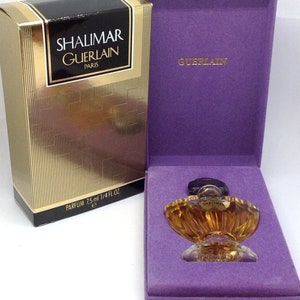 Può includere: Flacone di profumo Shalimar Guerlain in una scatola dorata con dettagli neri. Il flacone di profumo color ambra è esposto in una scatola foderata in velluto viola. La scatola recita "PARFUM 7,5 ml 1/4 FL OZ."