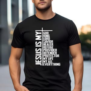 Könnte beinhalten: Schwarzes T-Shirt mit dem weißen Schriftzug "JESUS IS MY" auf der linken Seite und einer Liste von Wörtern wie "GOD", "KING" und "EVERYTHING". Das Shirt hat einen Rundhalsausschnitt und kurze Ärmel.