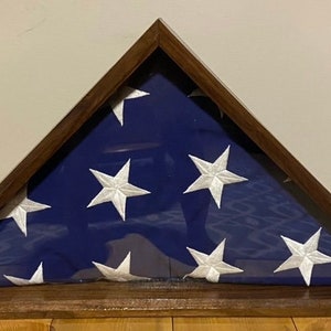 May include: A wooden flag display case with a folded American flag inside. The case is made of dark wood and has a glass front. The flag is folded in a triangular shape and is displayed in a traditional way.