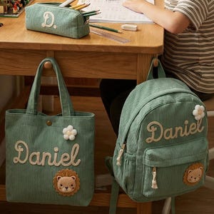 Könnte beinhalten: Ein Kinderschreibtisch mit einer weißen Schreibtischlampe, einem Federmäppchen, einer Tragetasche und einem Rucksack. Die grüne Cord-Tragetasche und der Rucksack sind mit dem Namen "Daniel" in Schreibschrift und einem Löwenaufnäher personalisiert. Das Federmäppchen hat den Buchstaben "D".