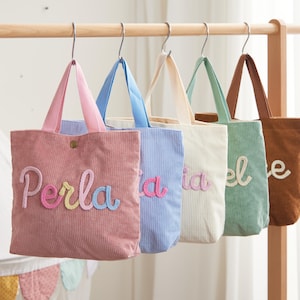 Pode incluir: Cinco sacolas de lona de veludo cotelê rosa, azul, creme, verde e marrom penduradas em um suporte de madeira. Cada sacola tem uma alça de cor diferente e um nome bordado em uma fonte divertida. A primeira sacola diz "Perla".