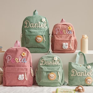 Könnte beinhalten: Fünf Rucksäcke und eine Tragetasche aus Cord in Rosa- und Grüntönen. Jede Tasche hat einen gestickten Namen, eine Blume und eine Tierapplikation. Die Taschen sind für Kinder konzipiert.