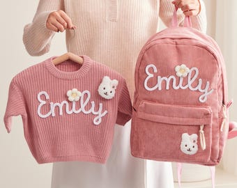 May include: A pink corduroy backpack and sweater set. The backpack and sweater are embroidered with the name "Emily" in white script. Both items feature a flower and a bear applique.