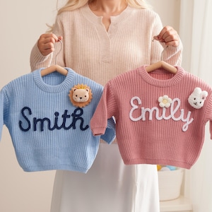 Könnte beinhalten: Zwei Baby-Pullover, einer hellblau und einer rosa, werden auf Holzbügeln präsentiert. Der blaue Pullover hat den Namen "Smith" in Dunkelblau gestickt und ein Löwen-Applikation. Der rosa Pullover hat den Namen "Emily" und eine Blumen- und Hasen-Applikation.
