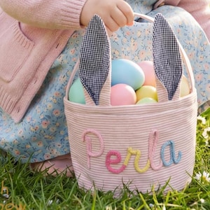 Könnte beinhalten: Ein rosa-weiß gestreifter Osterkorb mit Hasenohren, gefüllt mit pastellfarbenen Eiern. Der Korb hat den Namen "Perla" auf der Vorderseite gestickt. Die Szene spielt sich im Freien ab, mit einem Kind in einem rosa Cardigan und einem Blumenkleid.