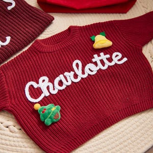May include: A red knitted sweater with the name "Charlotte" in white script. The sweater has a yellow bell and a green Christmas tree applique. A matching red hat and a piece of a red knitted item are in the background.