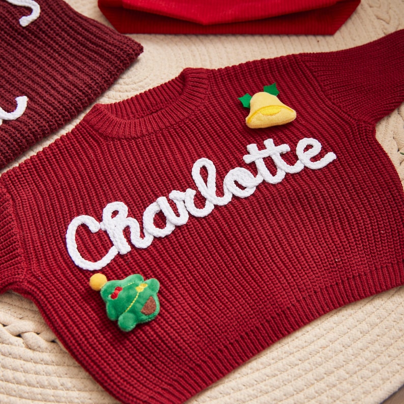 May include: A red knitted sweater with the name "Charlotte" in white script. The sweater has a yellow bell and a green Christmas tree applique. A matching red hat and a piece of a red knitted item are in the background.