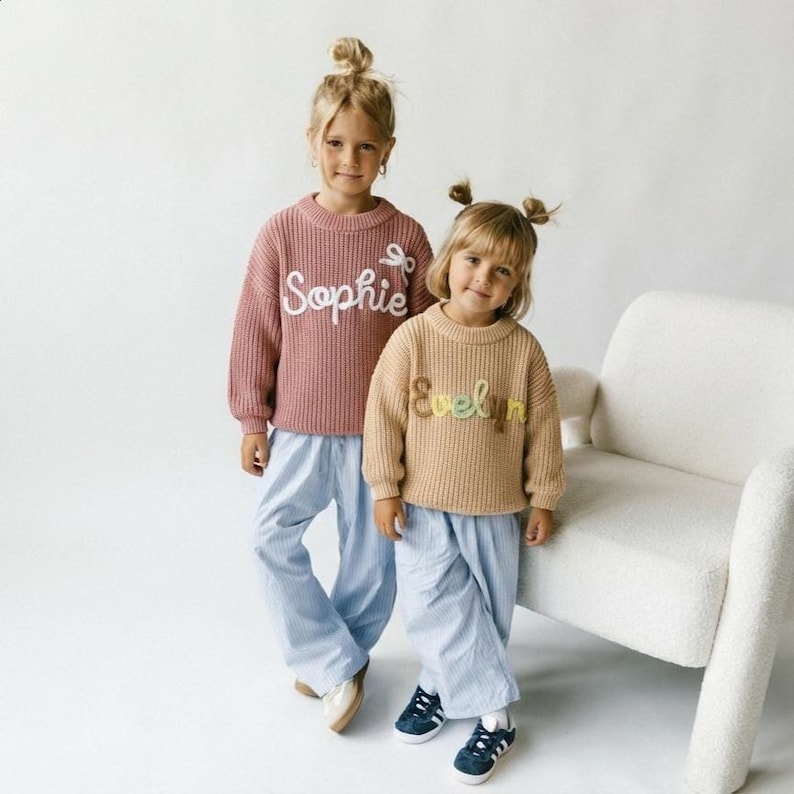 Op de afbeelding: Een oudroze gebreide trui met de wit geborduurde naam Sophie en een klein strikje, gecombineerd met een lichtblauwe gestreepte broek. Daarnaast een beige gebreide trui met de meerkleurig geborduurde naam Evelyn, gecombineerd met een lichtblauwe gestreepte broek.