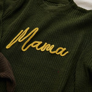 May include: Close-up of a dark green knit sweater with the word "Mama" embroidered in yellow cursive script. The sweater has a crew neck and a textured knit pattern. A portion of a brown sweater is visible.