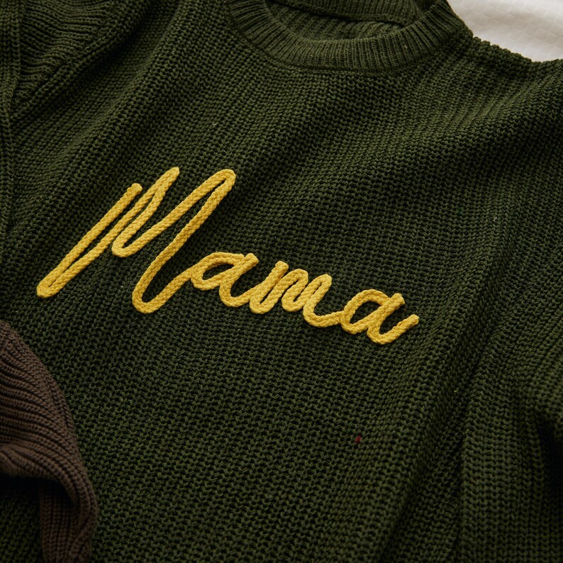 May include: Close-up of a dark green knit sweater with the word "Mama" embroidered in yellow cursive script. The sweater has a crew neck and a textured knit pattern. A portion of a brown sweater is visible.