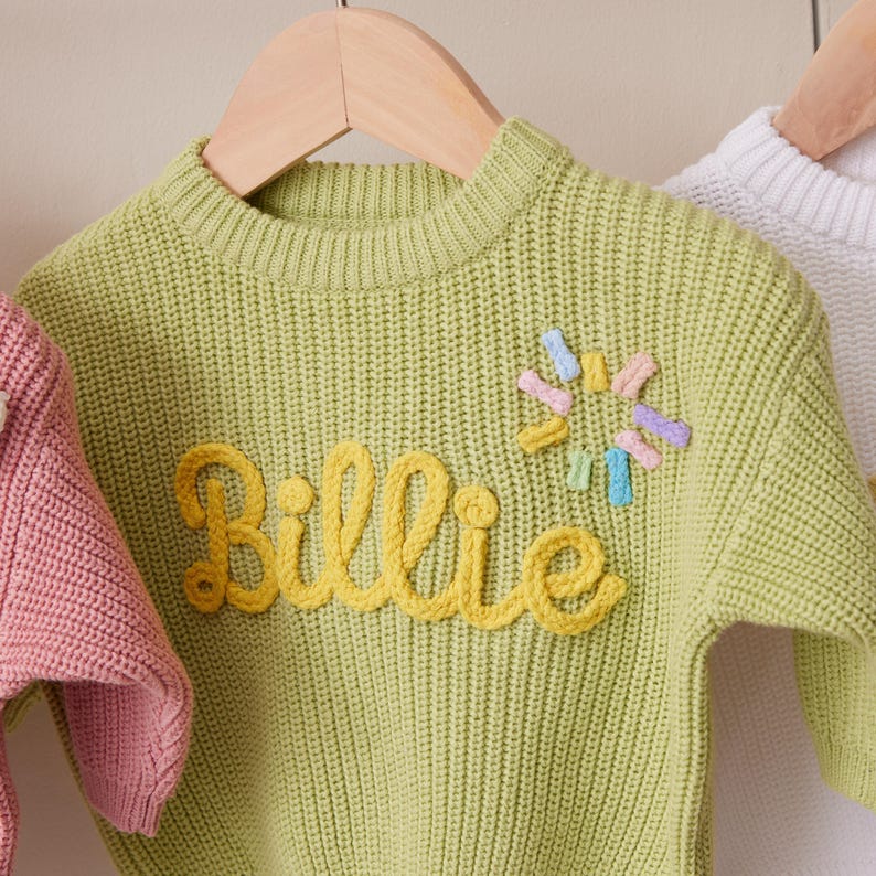 Op de afbeelding: Een limegroene gebreide trui met lange mouwen en de naam Billie in geel garen op de borst geborduurd. Rechts van de naam bevindt zich een klein, cirkelvormig zonachtig ontwerp gemaakt van korte, veelkleurige pasteltinten garen.