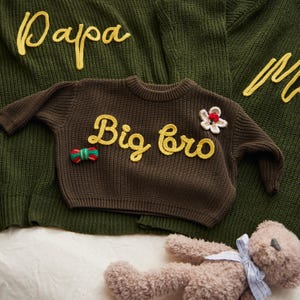 May include: A collection of knitted sweaters in olive green and brown. The brown sweater features the text "Big bro" in yellow, with a small flower and bow embellishment. The olive green sweaters have the words "Papa" and "Ma" in yellow. A teddy bear is in the foreground.