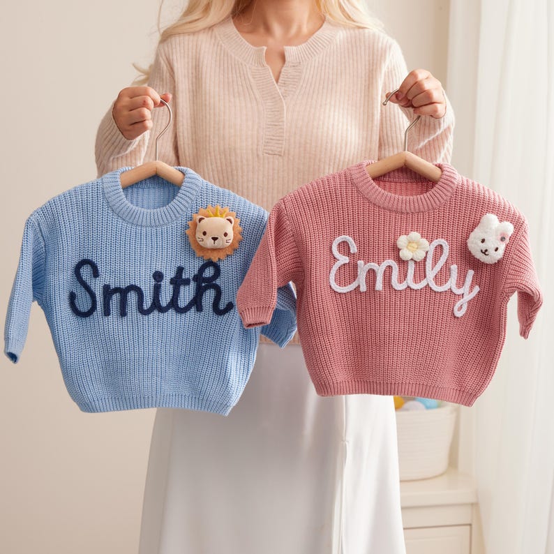 Op de afbeelding: Een lichtblauwe gebreide trui met de donkerblauw geborduurde naam Smith en een leeuwenpatch, en een roze gebreide trui met de wit geborduurde naam Emily, een bloemapplicatie en een konijnenpatch, beide op houten kledinghangers.
