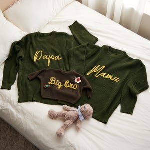 May include: Green knit sweaters with yellow embroidered text reading "Papa" and "Mama". A smaller brown sweater says "Big bro". A teddy bear lies nearby. The sweaters are on a white bed.